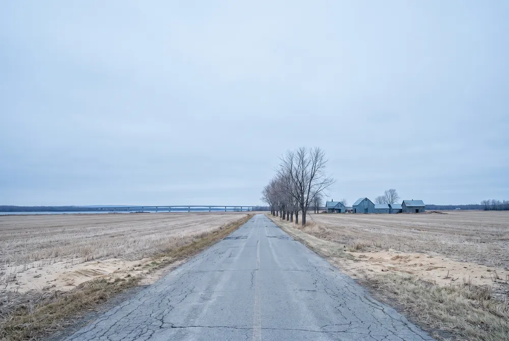 Illustration générée : une route droite entre des champs moissonnés sur la rive sud d'un large fleuve, des arbres nus et des bâtiments de ferme, un long pont bas à l'horizon